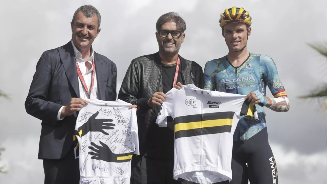 Javier Guillén, a la izquierda, y un representante de la marca de ropa ciclista Santini, entregan el maillot solidario a Luis León Sánchez, que se retiró tras La Vuelta 2023
