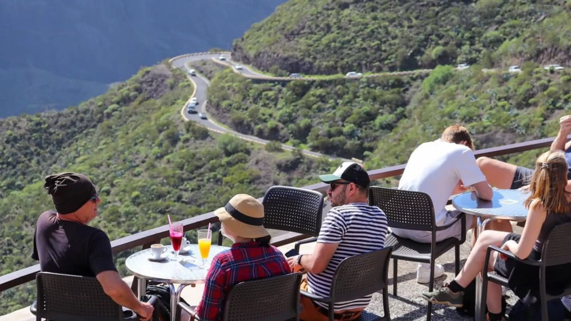 Unas personas en la cafetería del mirador Cruz de Hilda del Parque Rural de Teno. De fondo, la carretera estrecha y serpenteante de Masca, con tráfico./ CAFETERÍA MIRADOR CRUZ DE HILDA
