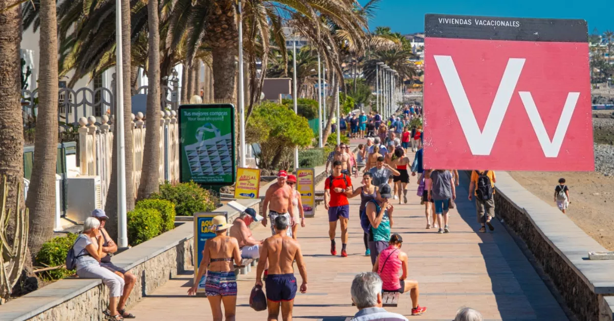 Varias personas paseando en Gran Canaria junto al cartel de Viviendas Vacacionales. / MONTAJE AH