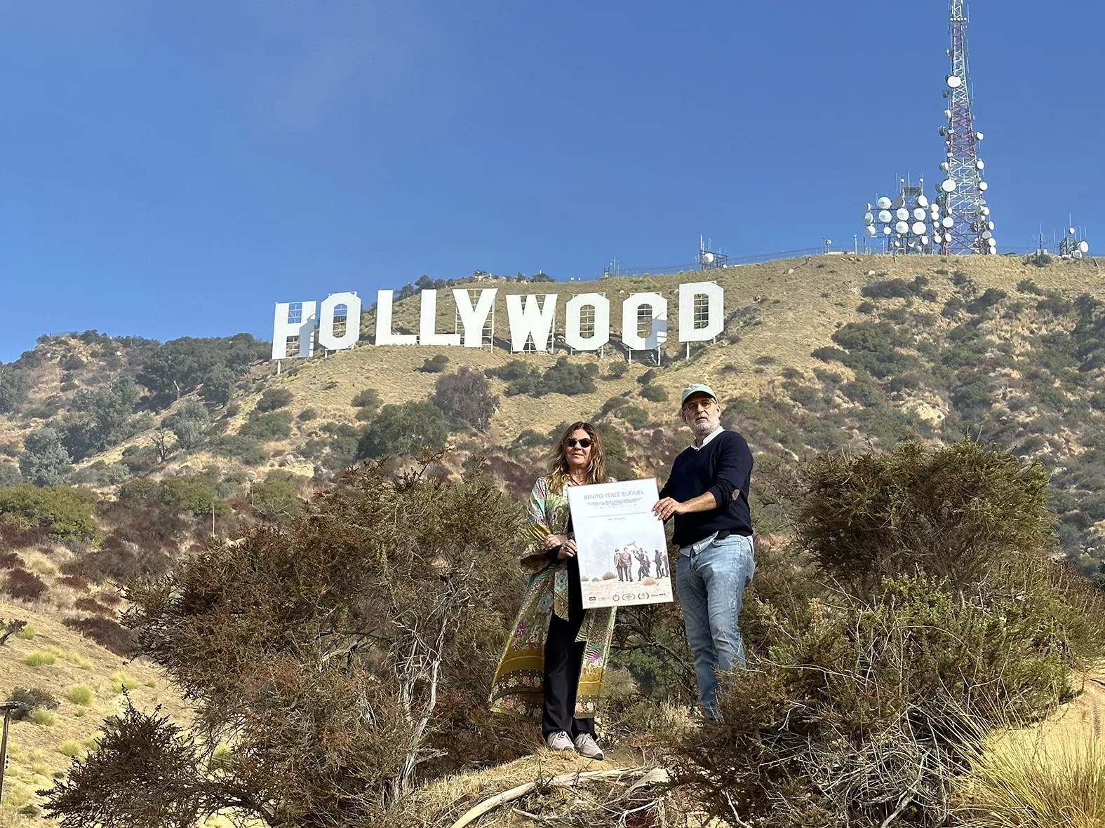Marta de Santa Ana, productora del filme, y Luis Roca, director de la película, sostienen el cartel de 'Benito Pérez Buñuel' delante del cartel de Hollywood en Los Ángeles. / AH
